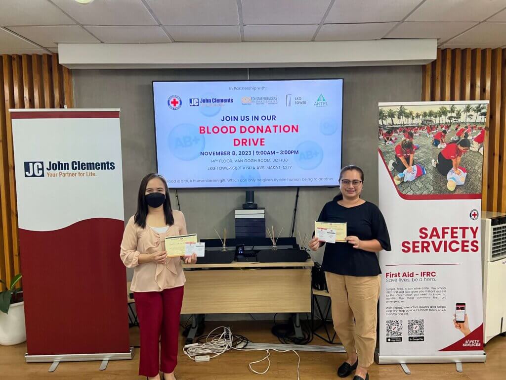 two women holding up papers and standing with a TV screen bearing blood donation drive